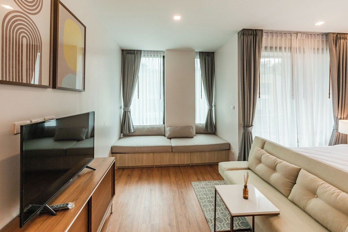 A modern hotel room with a large window, a sofa, a TV, and a small table.