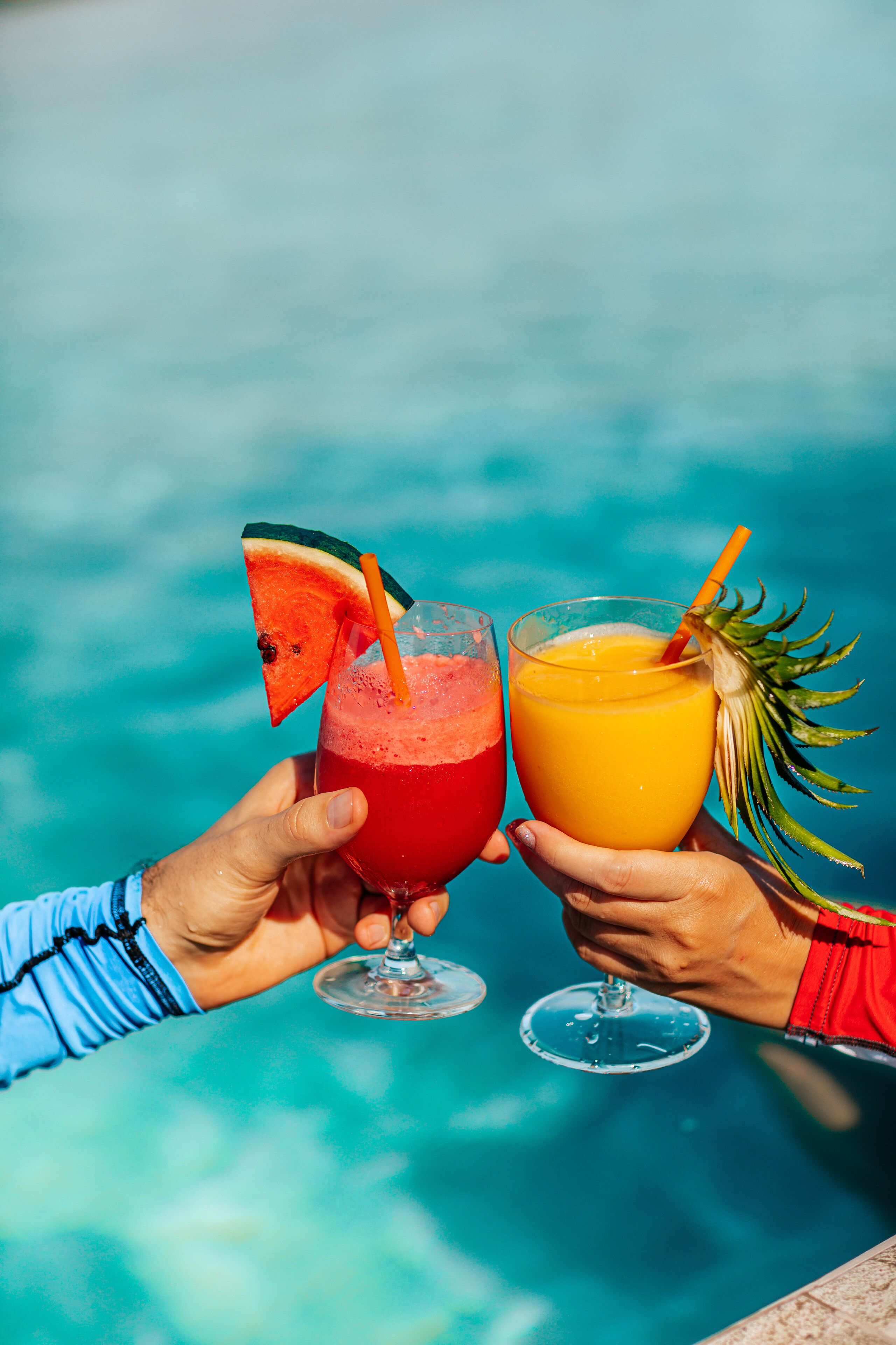Zwei Hände halten Gläser mit Getränken, eines mit Wassermelone und das andere mit tropischer Dekoration, vor blauem Hintergrund.