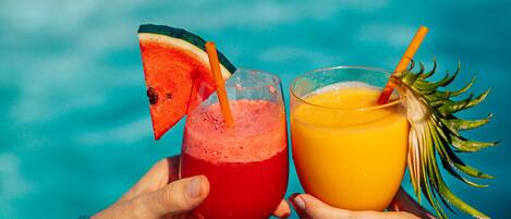 Dos manos sosteniendo vasos con bebidas, uno con una tajada de sandía y otro con un adorno tropical, sobre un fondo azul.