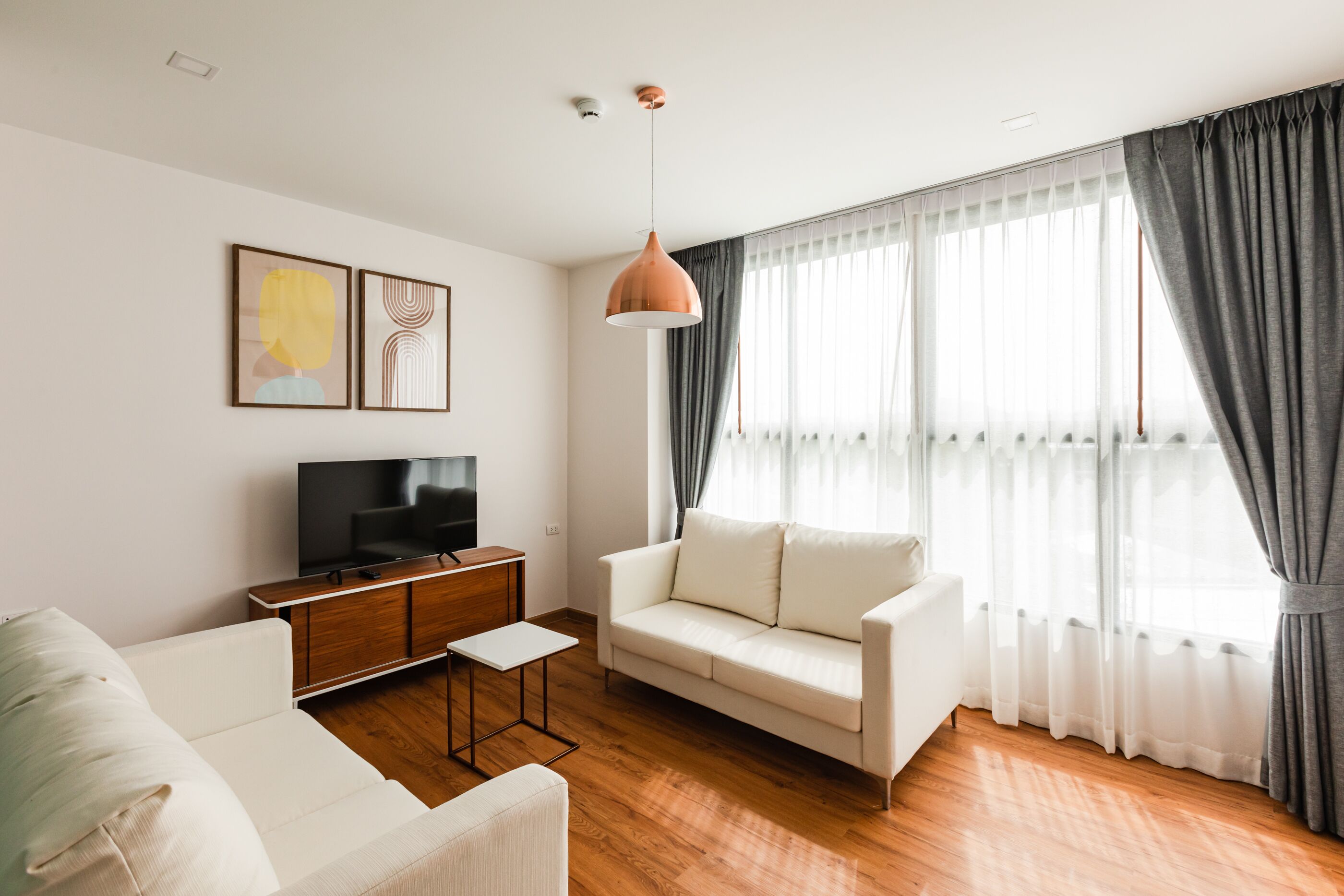 A modern living room with a white sofa, a wooden coffee table, a TV on a wooden cabinet, and a large window with grey curtains.