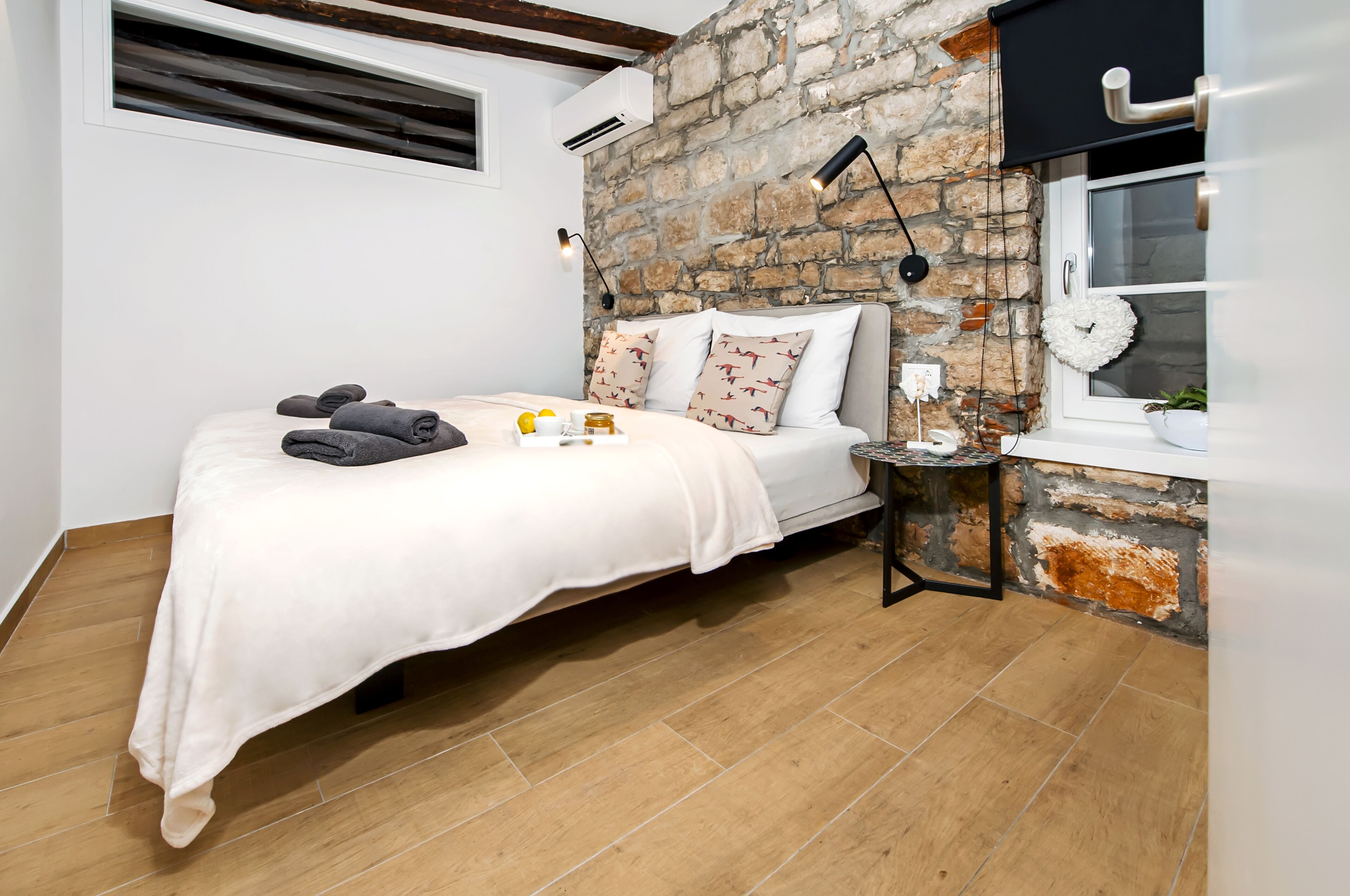 A bedroom with a stone wall, a bed with a white bedspread, two pillows, a bedside table with a lamp, and a window with a wreath.