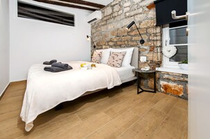 A bedroom with a stone wall, a bed with a white bedspread, two pillows, a bedside table with a lamp, and a window with a wreath.