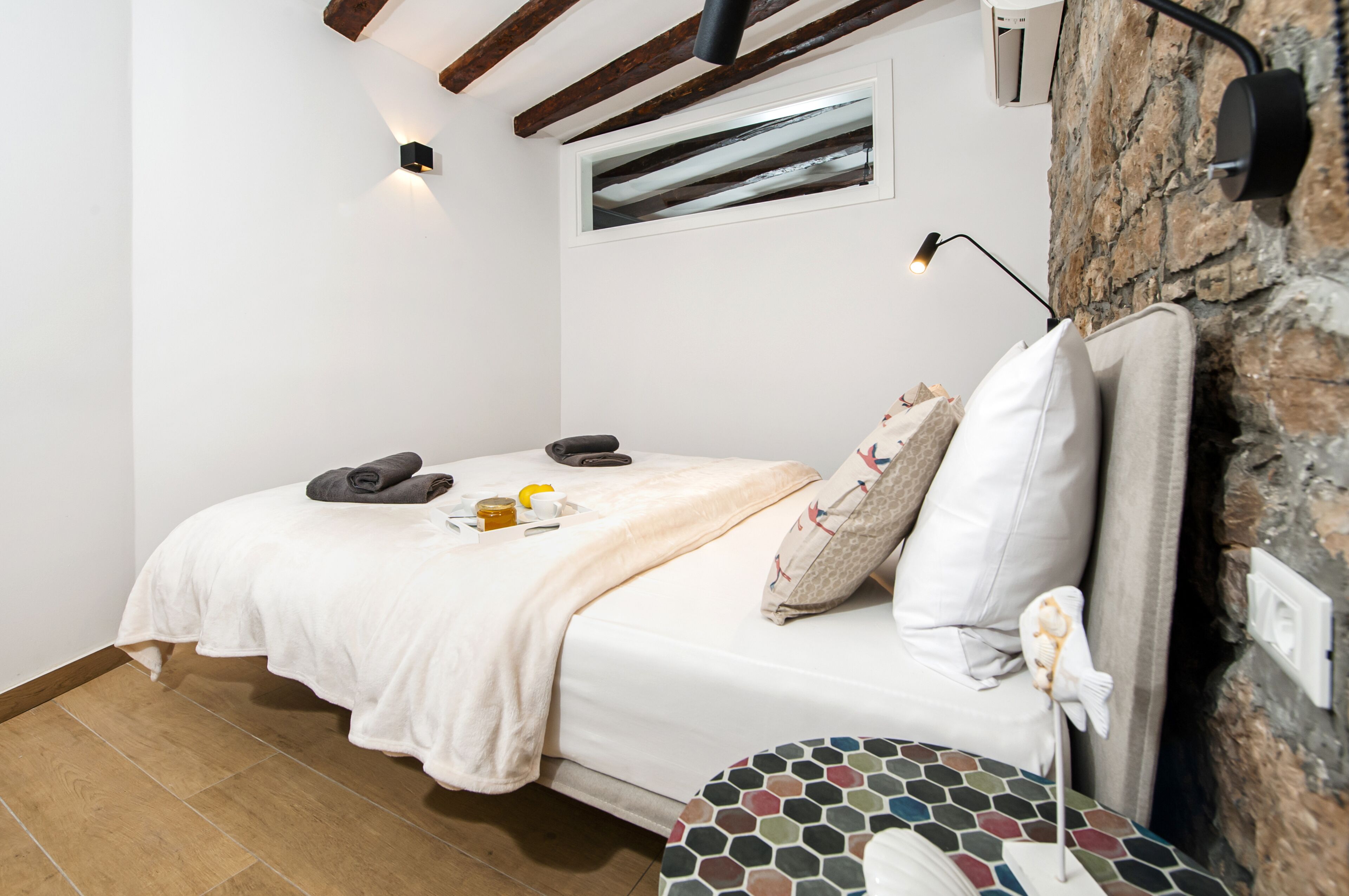 A bedroom with a bed, a small table with a decorative item, a wall-mounted light, and a stone wall.