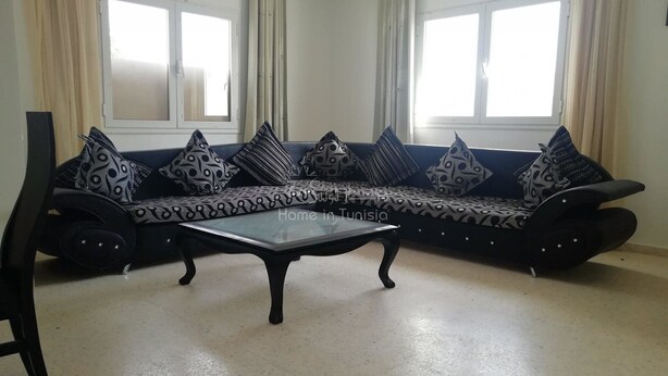 A living room with a black sofa adorned with patterned cushions, a glass coffee table, and a view of a window with curtains.