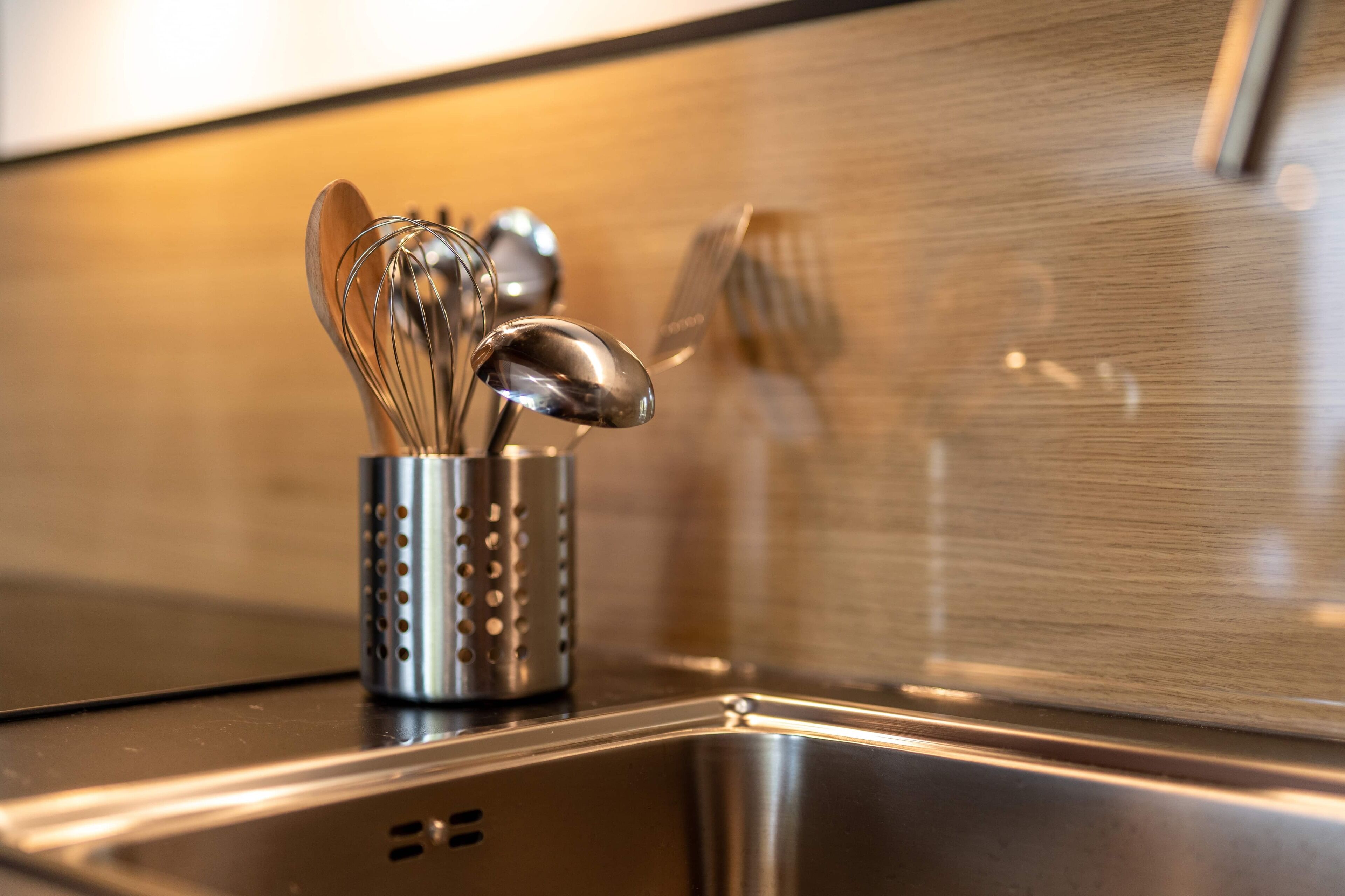 A stainless steel kitchen sink with a utensil holder containing a whisk, spatula, and spoon.