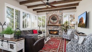 A living room with a fireplace, a stone wall, a ceiling fan, and a large window with blinds. - Hill-top Haven w/ Heated pool & spa, expansive views and more! (Sedona)