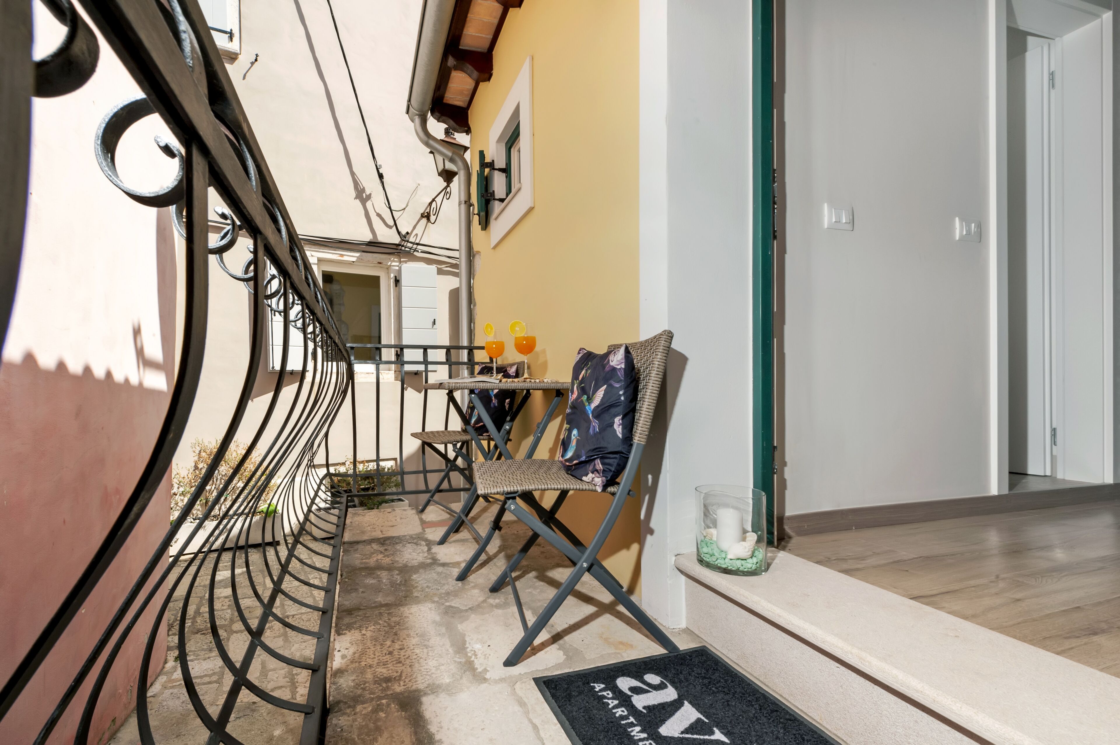 Een balkon met een metalen hekwerk, een tafel met stoelen en een deur aan de rechterkant.
