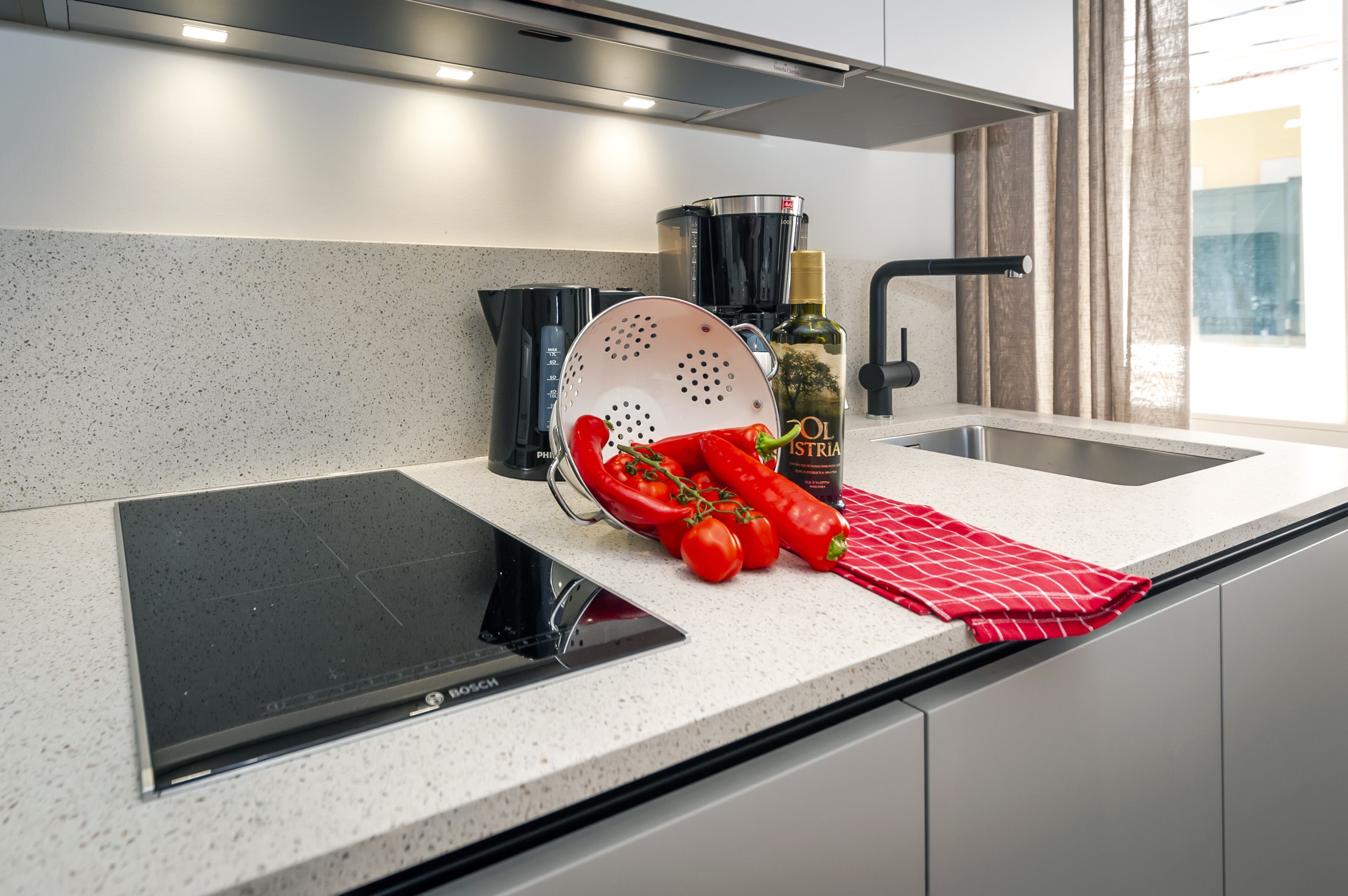 A modern kitchen with a Bosch induction cooktop, a coffee maker, a colander, red chili peppers, and a bottle of olive oil.