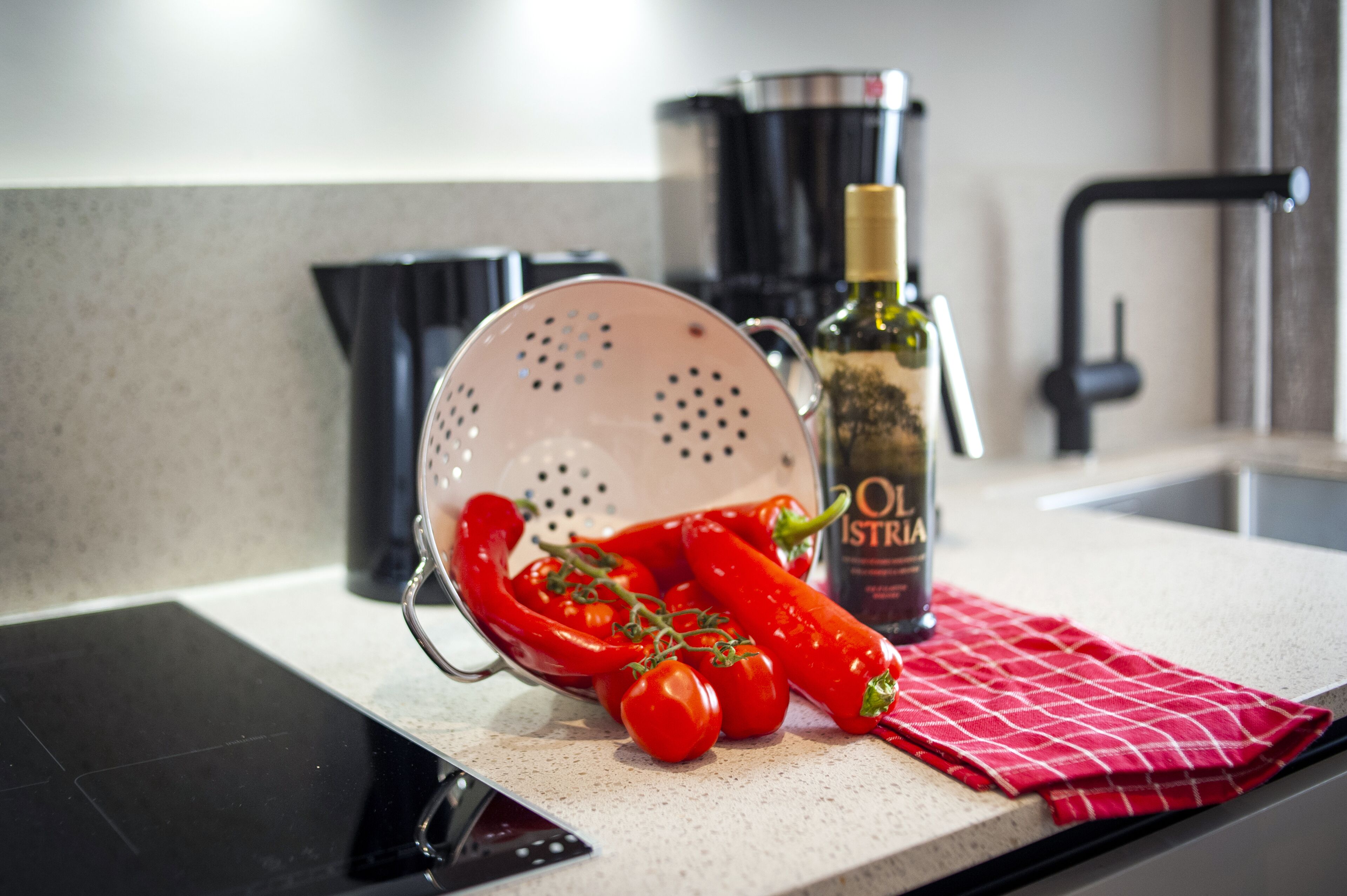 A kitchen counter with a colander, red chili peppers, a bottle of olive oil, and a red checkered cloth.