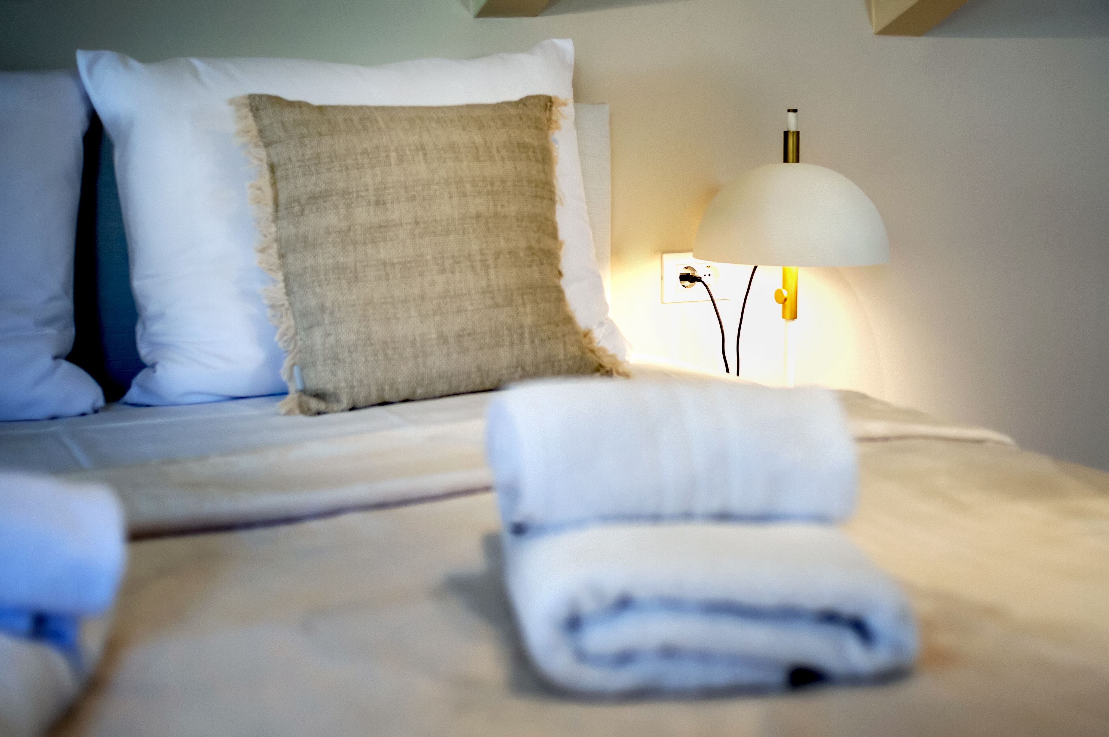 Una cama bien tendida con sábanas blancas y una toalla doblada, un cojín beige y una lámpara blanca sobre una mesita de noche.