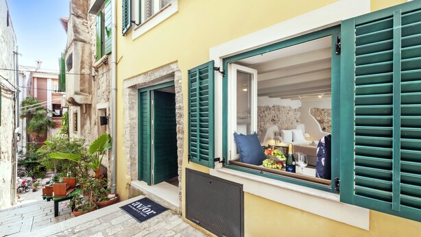 A narrow street with a yellow building featuring a window with green shutters, a stone wall, and a small outdoor seating area with plants. - Yellow House Rovinj (Rovinj)
