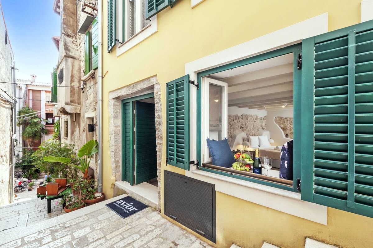 A narrow street with a yellow building featuring a window with green shutters, a stone wall, and a small outdoor seating area with plants.