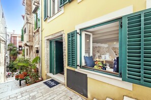 Una calle estrecha con un edificio amarillo que presenta una ventana con contraventanas verdes, un muro de piedra y un pequeño área exterior con plantas.