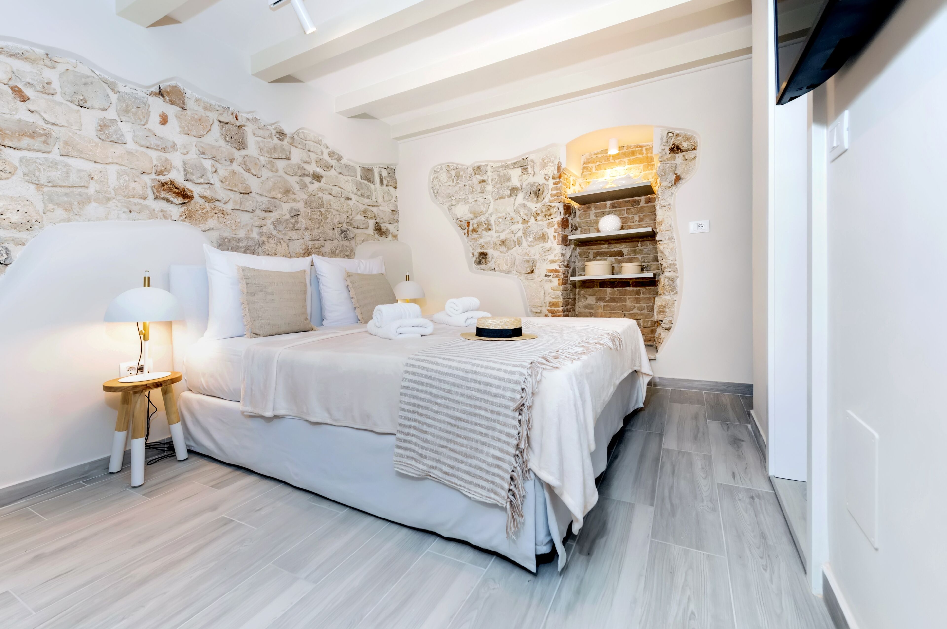 Un dormitorio con pared de piedra, una cama con ropa blanca, una mesita de noche con lámpara y un televisor montado en la pared.
