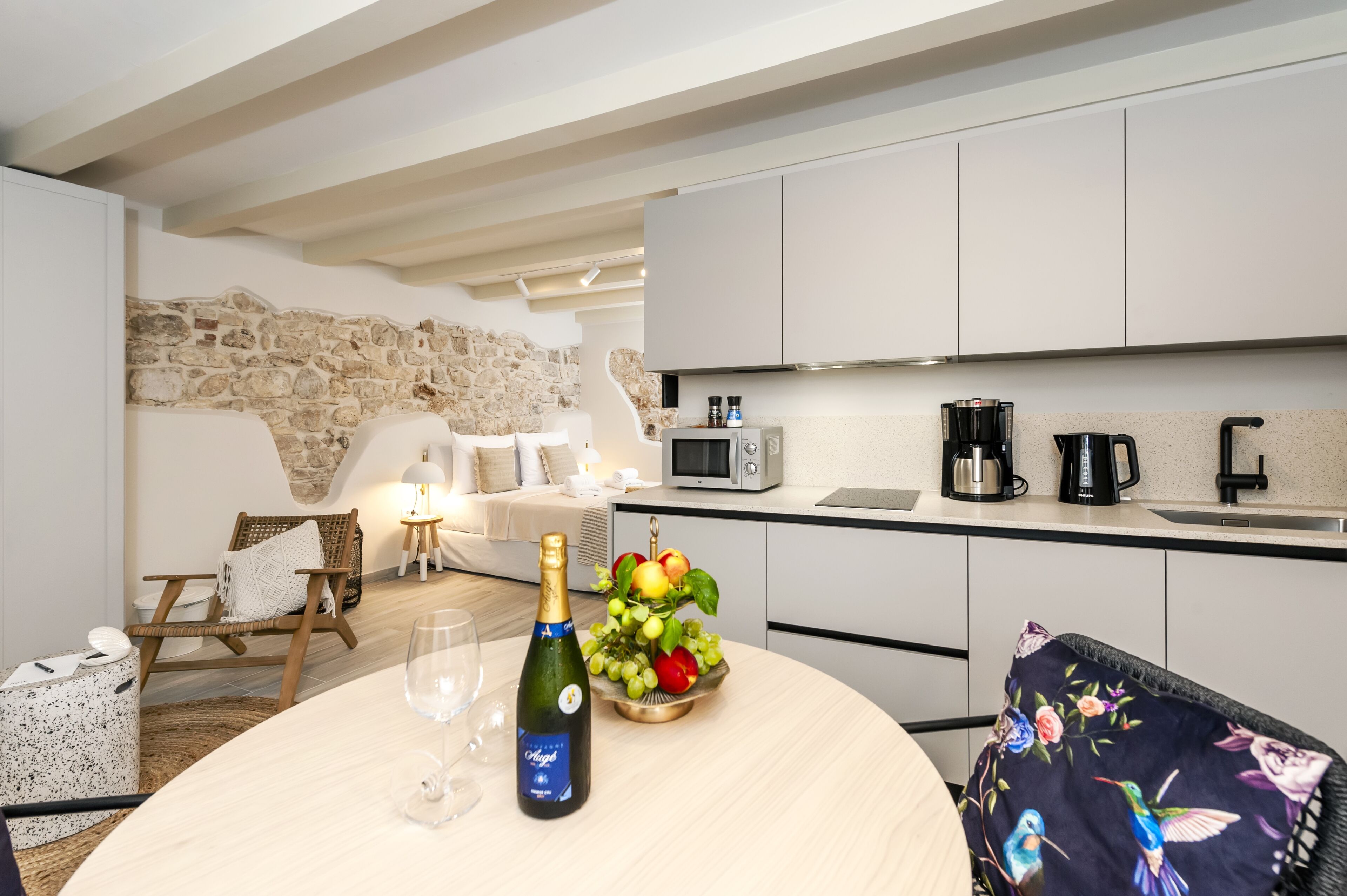 Una cocina moderna con armarios blancos, pared de piedra, zona de comedor con mesa redonda y una silla de mimbre.