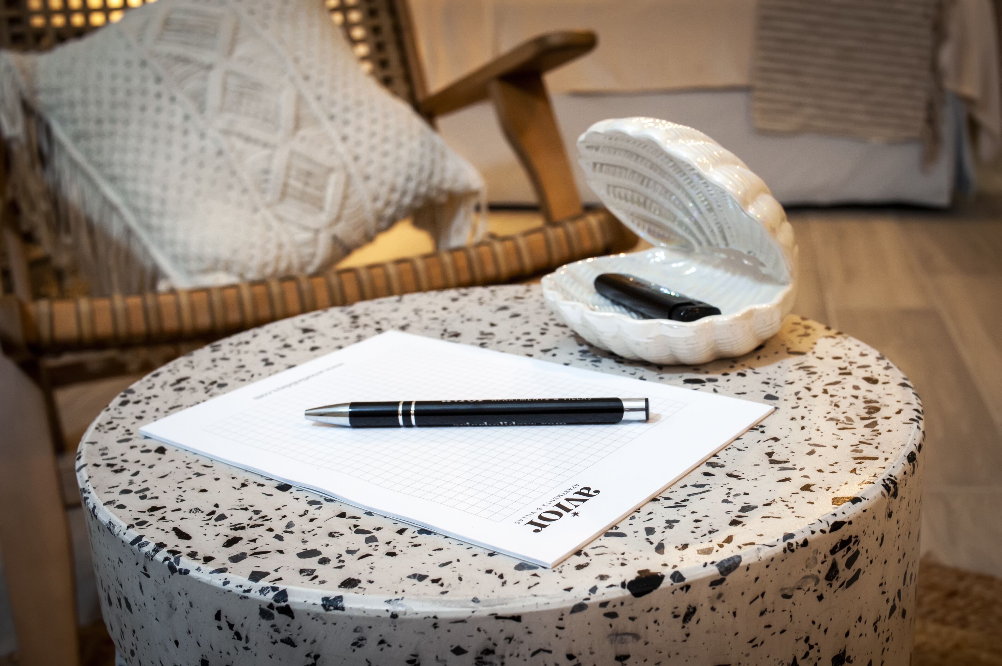 Ein runder Tisch mit Notizbuch und Stift, einer Keramiktasse und einem Korbstuhl im Hintergrund.