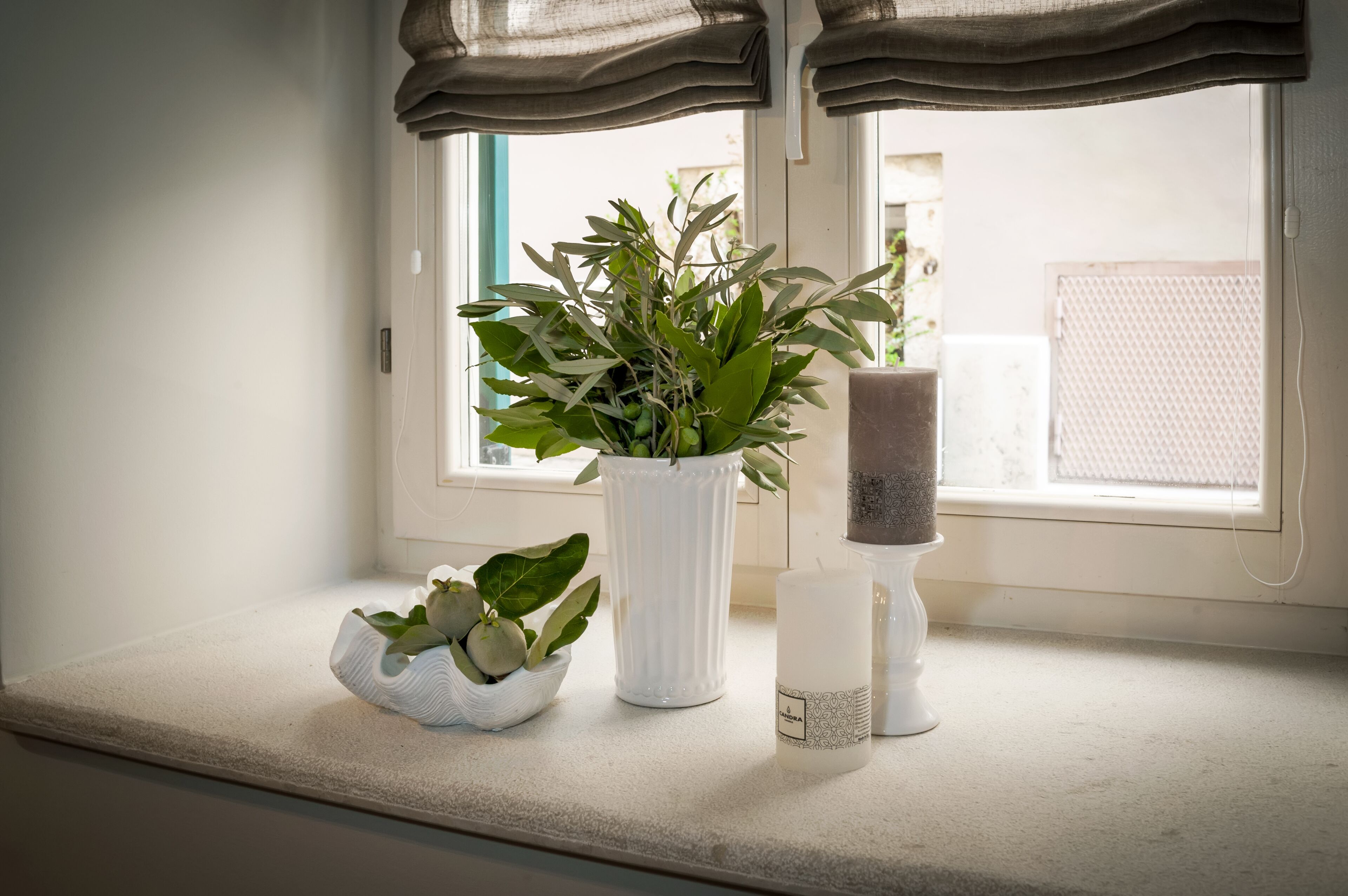 Un alféizar con un jarrón blanco que contiene plantas verdes, una vela blanca y un candelabro blanco.