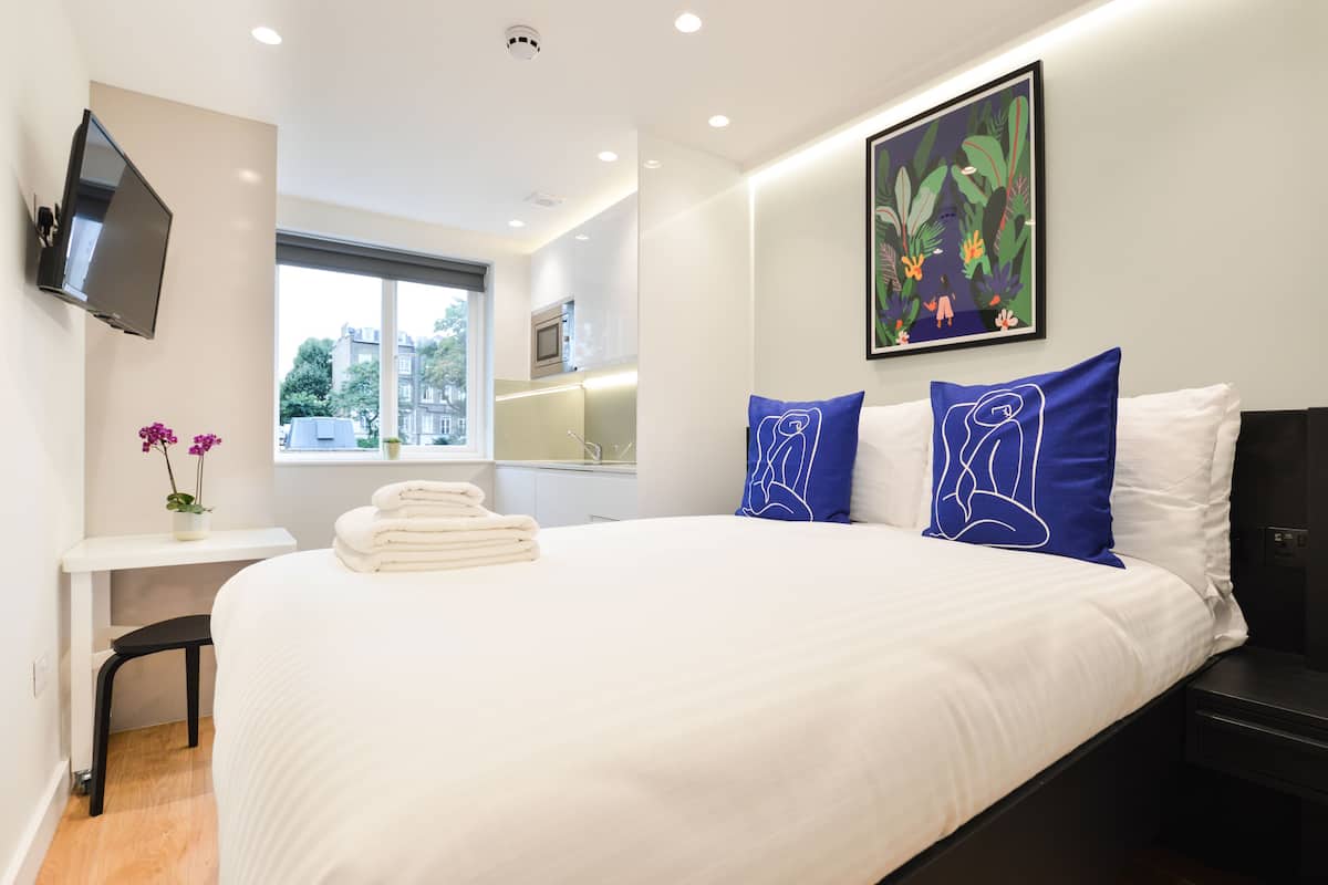 Ein modernes Hotelzimmer mit einem großen Bett, einem flachbildfernseher an der Wand, einem kleinen Schreibtisch mit Stuhl und einem Fenster mit Blick auf die Stadt.