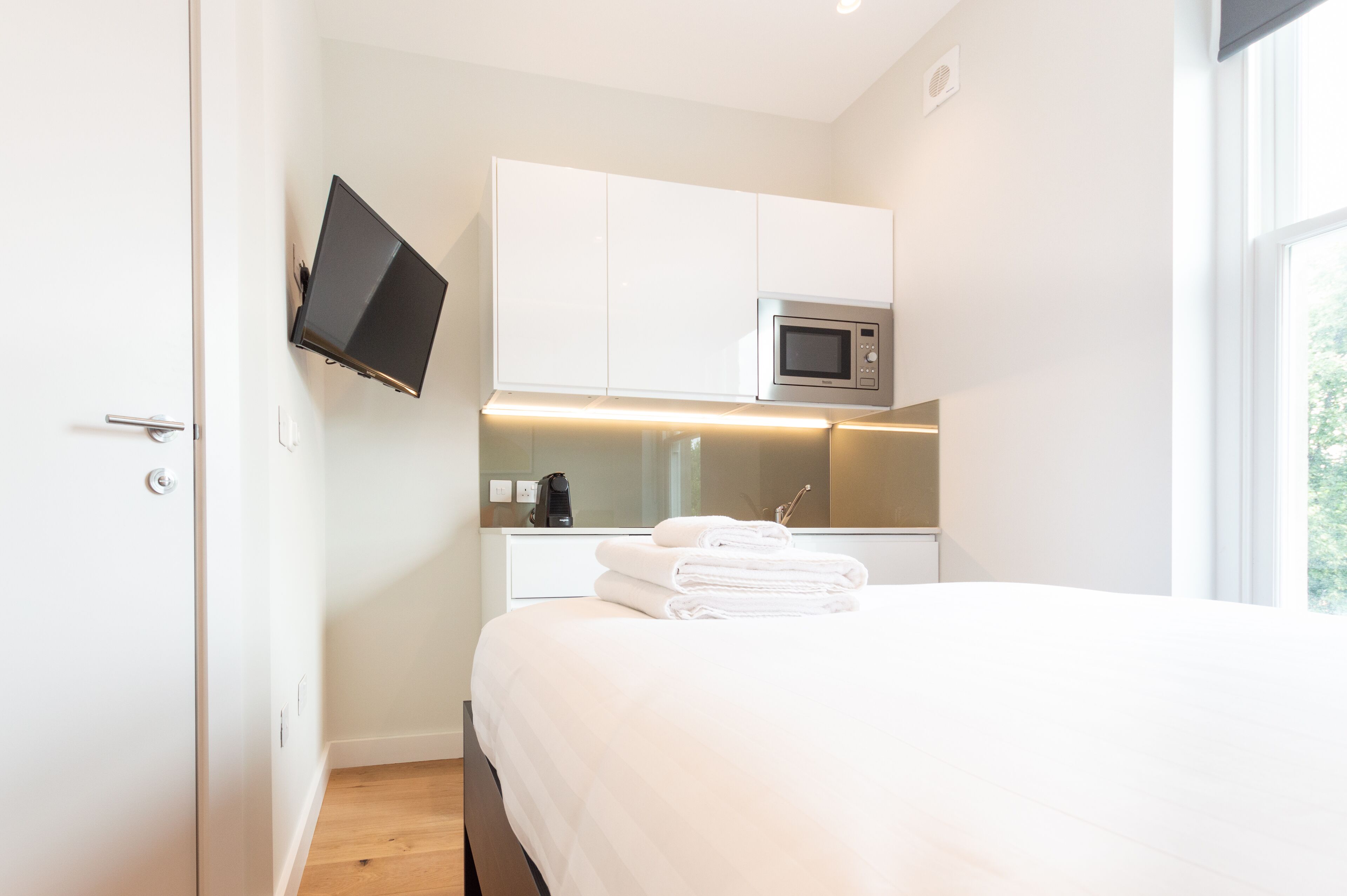 A modern hotel room with a bed, a flat-screen TV mounted on the wall, a microwave, and a small table with a coffee maker.