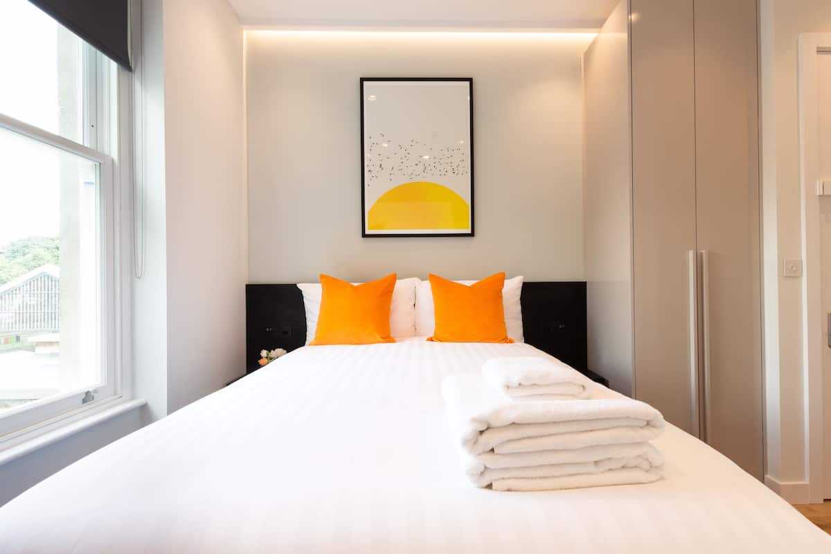 Ein ordentlich bezogenes Bett mit weißen Leinentextilien und orangefarbenen Kissen, ein gerahmtes Kunstwerk darüber und ein Blick ins Freie durch ein Fenster.