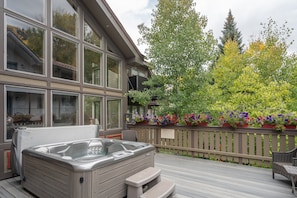 A hot tub on a wooden deck, a building with large windows, and a fence with flowering plants. - 4 BDR Mountain Retreat w/ private deck/hot tub/steps to trail (Telluride)