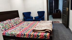 A neatly made bed with a colorful patterned bedspread and a folded blanket. There are two blue armchairs and a dark wooden headboard. A small rug is on the floor, and a door is visible in the background. - Private & Secure 1BHK Flat - Central location Ph7 (Rawalpindi)