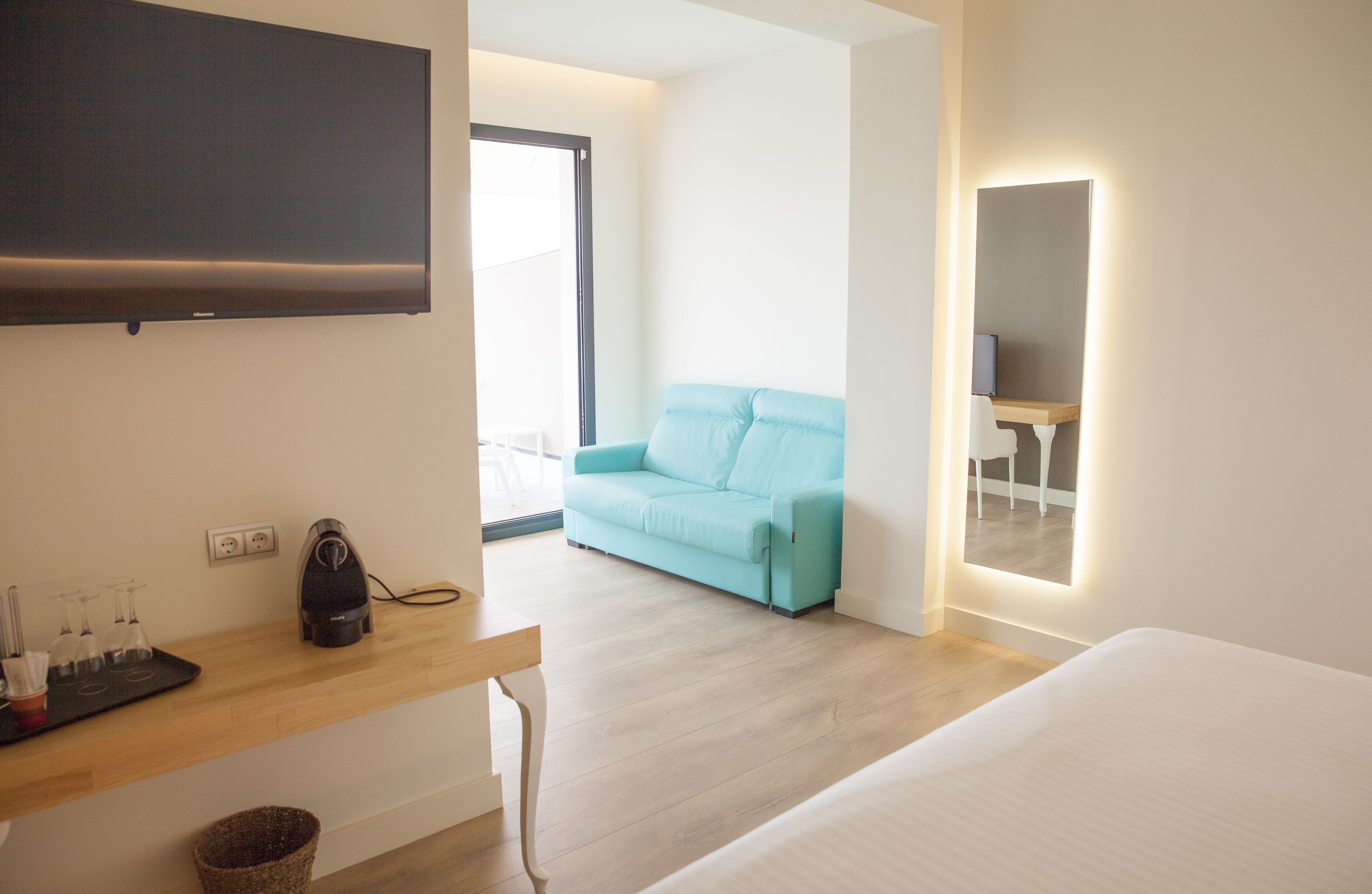 A modern hotel room with a flat-screen TV, a wooden desk with a kettle, a light blue sofa, and a bed.