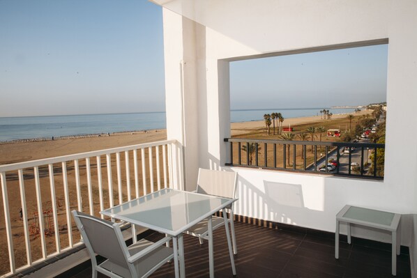 A balcony with a glass table and chairs, overlooking a beach with palm trees and a road with cars. - Hotel Brisamar Suites (El Vendrell)