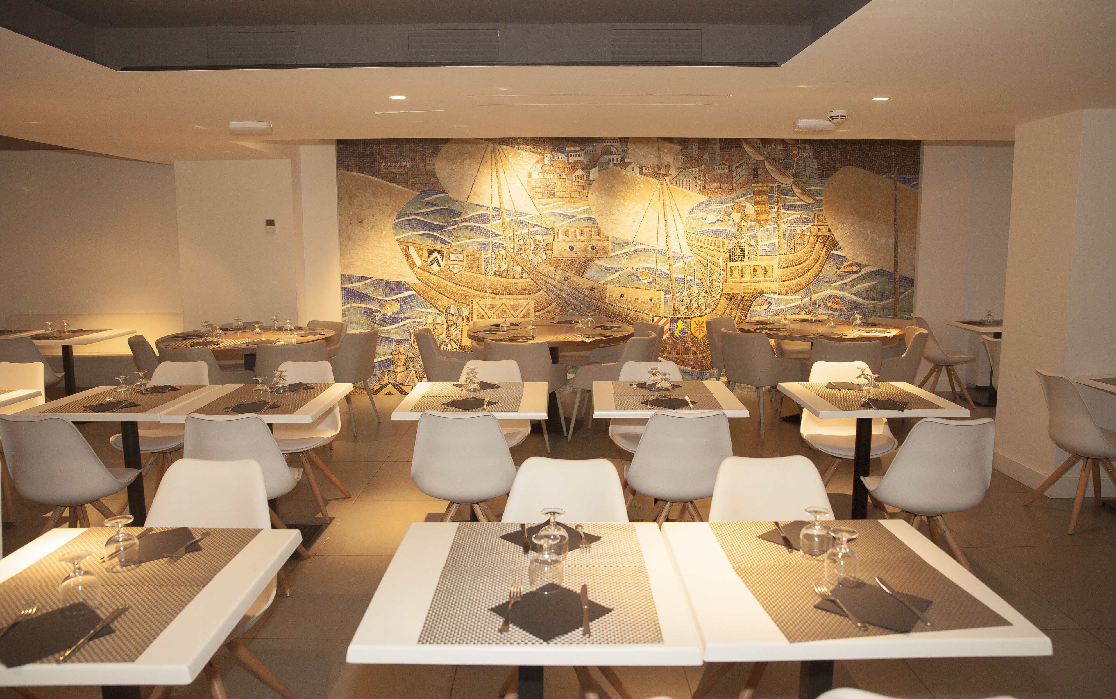 A restaurant with multiple tables and chairs, a mosaic wall, and a modern ceiling design.