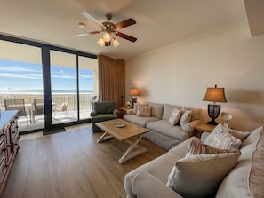 A spacious living room with a large sofa, a coffee table, and a ceiling fan. There is a balcony with a view of the beach. - Beautiful 3BR/2BR condo! (Dauphin Island)
