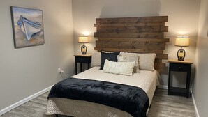 A bedroom with a bed, two bedside tables, a wall-mounted lamp, a painting, and a wooden headboard.