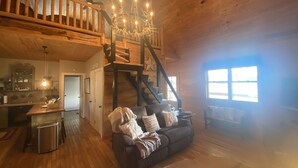 A rustic cabin interior with a wooden staircase, a cozy living area with a sofa, a kitchen area, and a large window providing natural light.