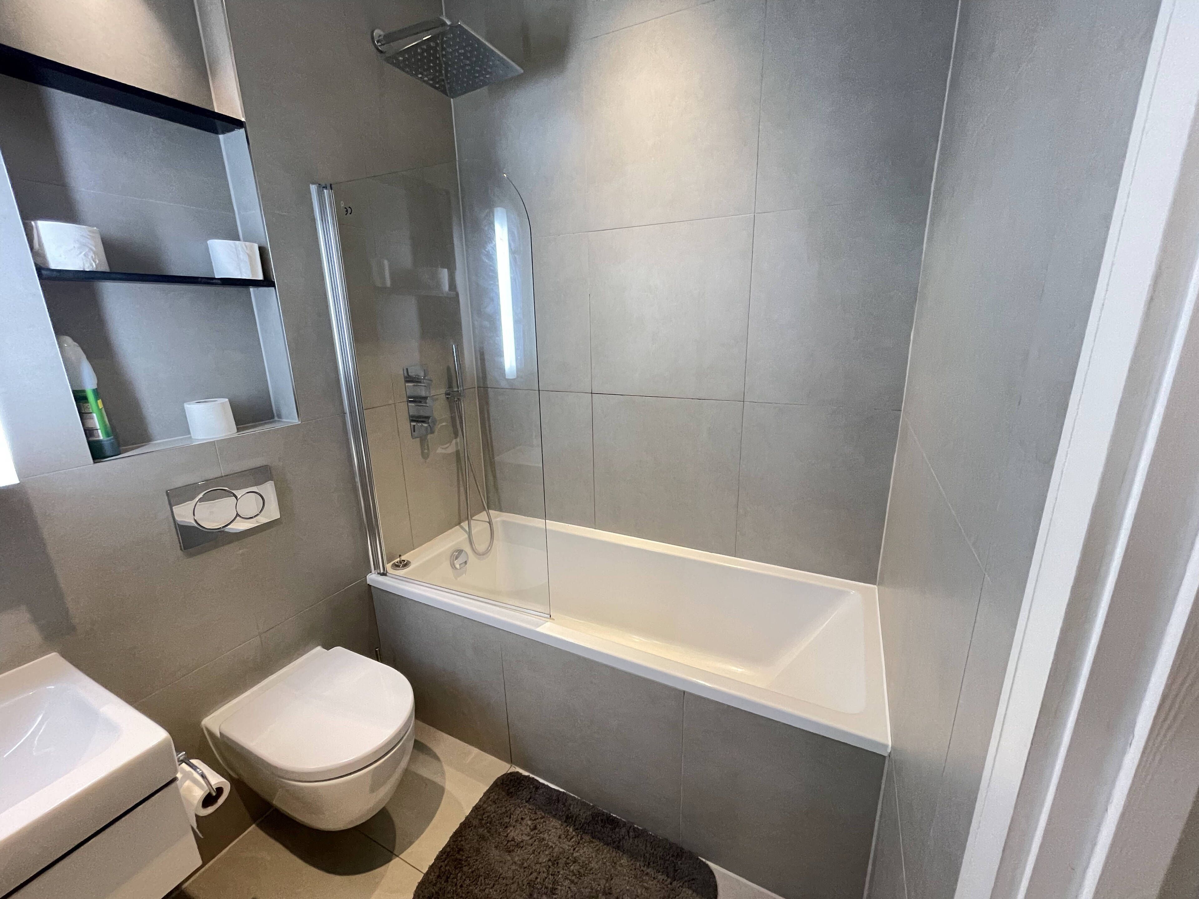 Un baño moderno con ducha de vidrio, una bañera blanca, inodoro y un espejo con repisas.