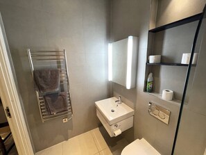 A modern bathroom with a towel rack, sink, mirror, and shelves with toiletries. - Stunning 1-bed Deluxe Apartment in Slough (Slough)