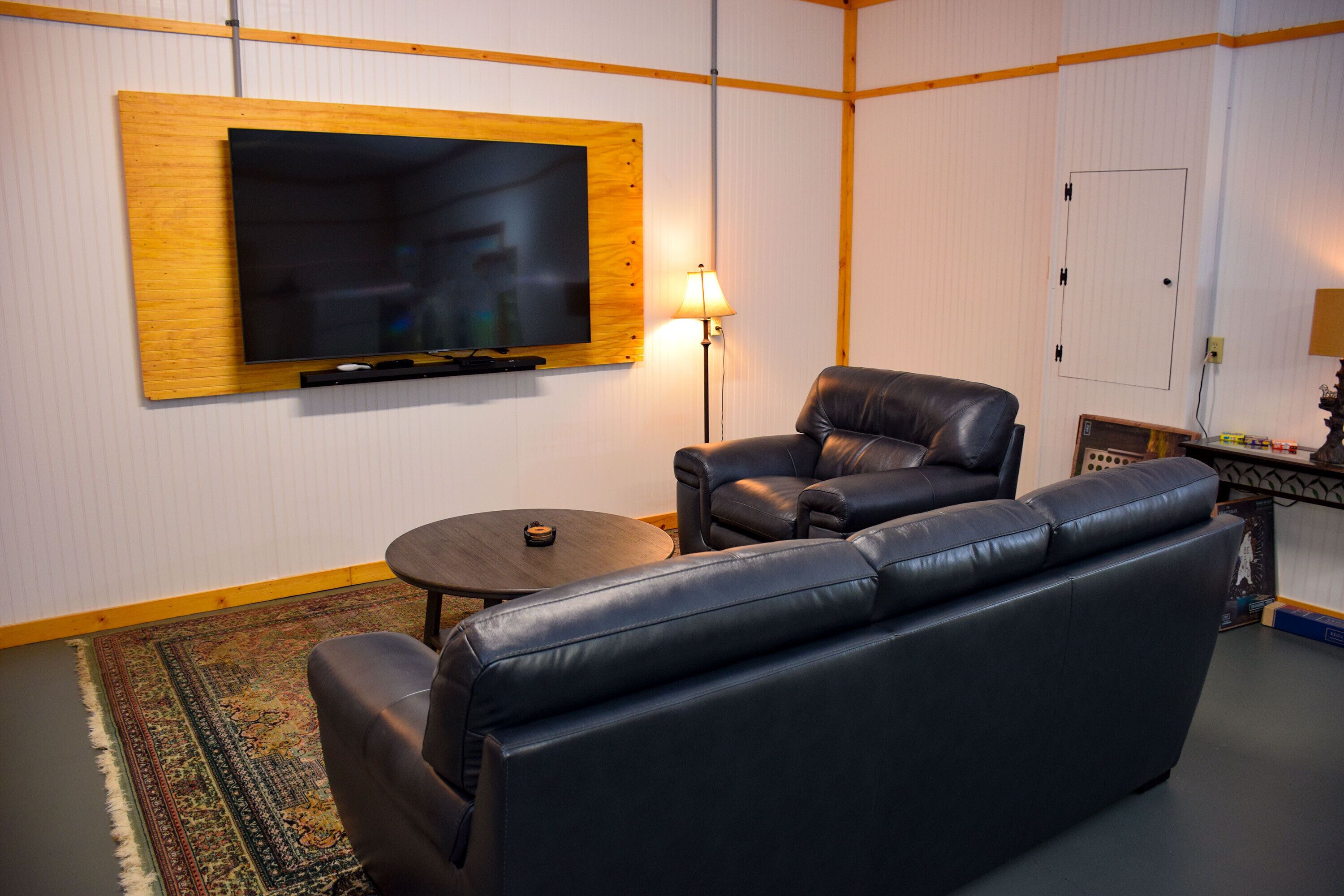 A living room with a large flat-screen TV mounted on a wooden panel, a leather sofa, a small round coffee table, and a lamp.