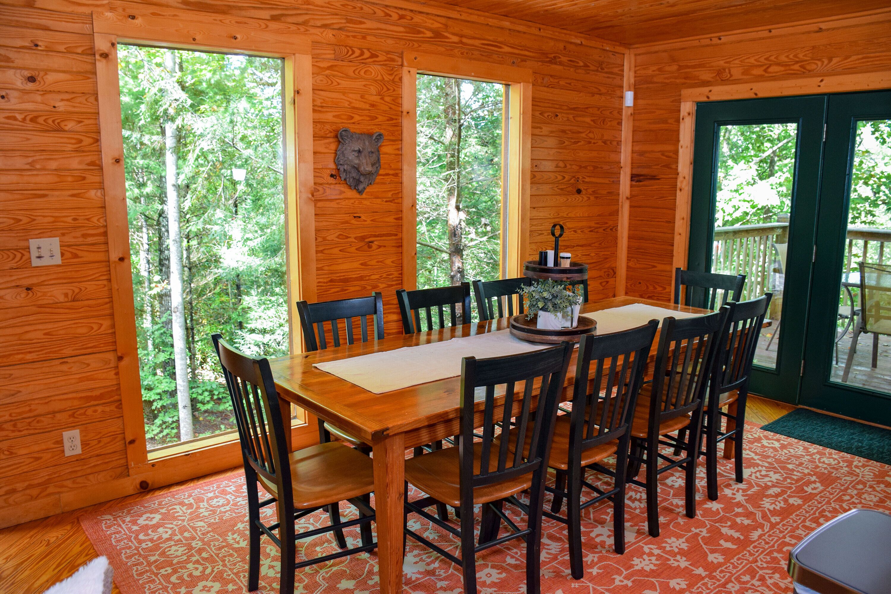 A wooden dining room with a large table and chairs, a bear head decoration, and a view of the forest through large windows.