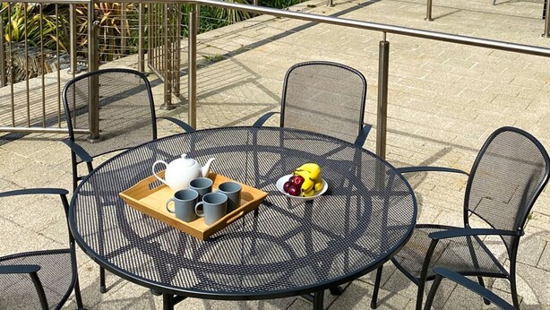 Un área de estar al aire libre con una mesa redonda y sillas, una bandeja con tazas y un plátano, y vista a la costa.