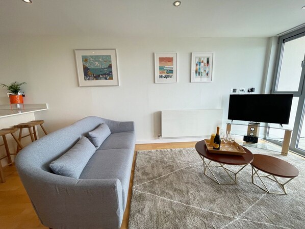 A modern living room with a grey sofa, a coffee table with a tray of drinks, a TV, and framed artwork on the wall. - 8 Zinc - an apartment that sleeps 4 guests in 2 bedrooms (Newquay)