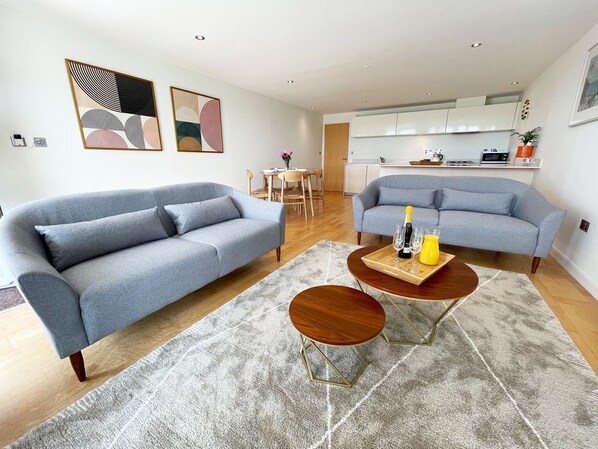 Un salon moderne avec deux canapés gris, une table basse en bois et une cuisine en arrière-plan.