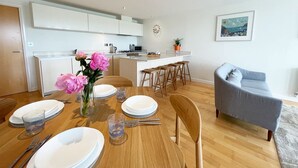 Una cocina moderna con armarios blancos, una zona de comedor con mesa de madera preparada para cuatro personas, y un sofá gris en la sala de estar.