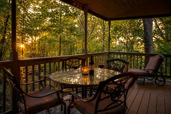A covered outdoor patio with a round table and chairs, surrounded by a wooden deck and a railing, overlooking a forested area.