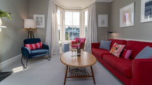 Un salon avec un canapé rouge, un fauteuil bleu, une table ronde en bois et une vue dégagée sur l’extérieur grâce à une grande baie vitrée.