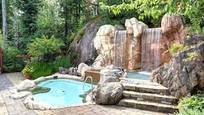 Ein naturbelassener Poolbereich mit Wasserfall, umgeben von Steinen und viel Grün.