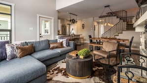 Ein geräumiges Wohnzimmer mit einem grauen Ecksofa, einem runden Couchtisch und einem Kamin aus Stein. Eine Treppe mit Metallgeländer führt zu einem Essbereich mit Holztischen und -stühlen.