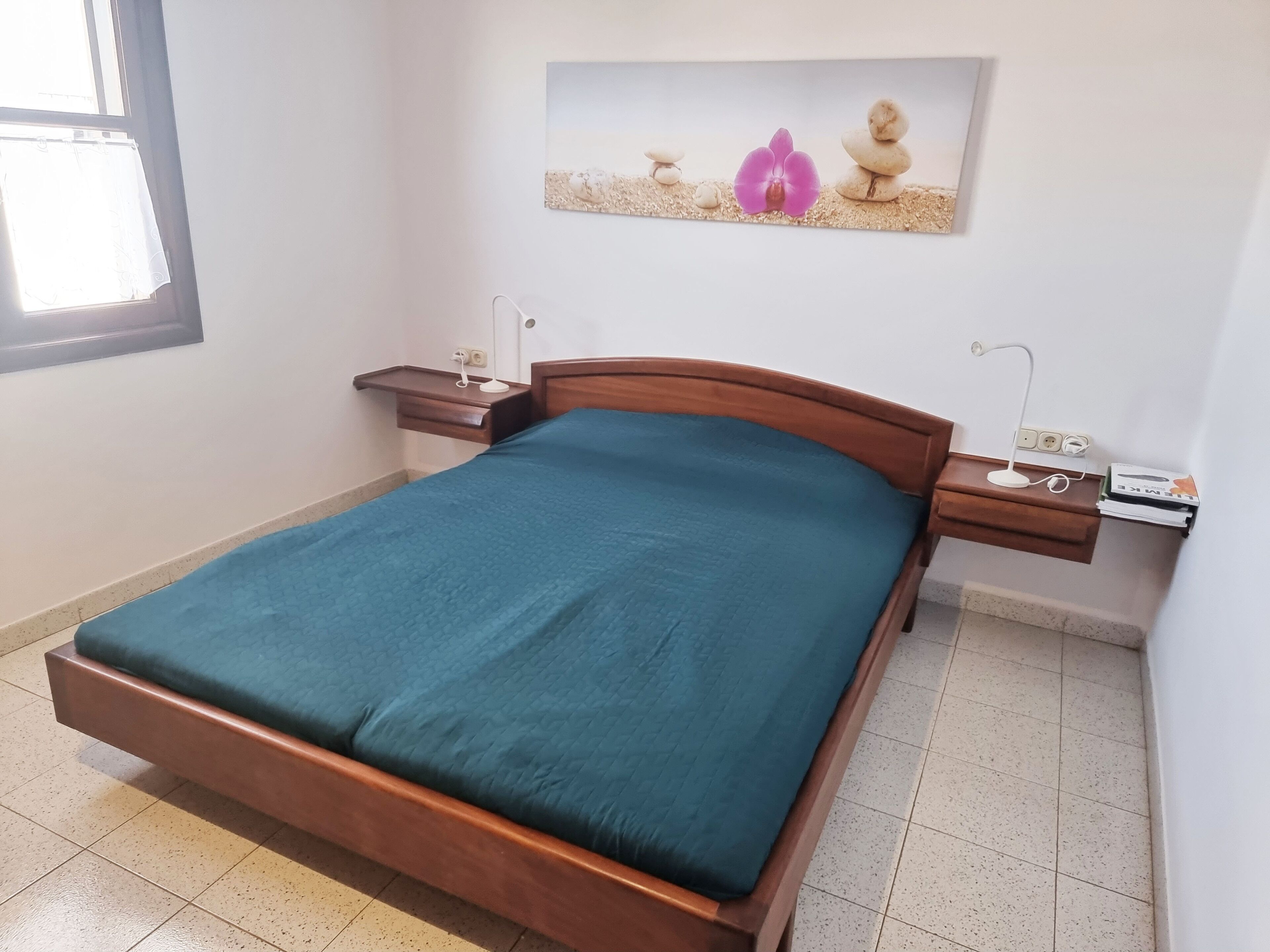 Un lit bien fait, recouvert d’une couvre-lit bleue, se trouve dans une pièce aux murs blancs. Une petite table de chevet, sur laquelle se trouve une lampe et une prise électrique, est placée à côté du lit. Un tableau encadré est accroché au mur, au-dessus du lit.