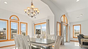 Un comedor amplio con una mesa grande y sillas blancas, una araña y ventanas grandes con marcos de madera.