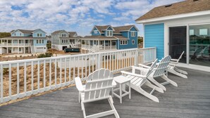 Una terraza de madera con sillas Adirondack blancas y una mesa, con vistas a una playa con varias casas azules.