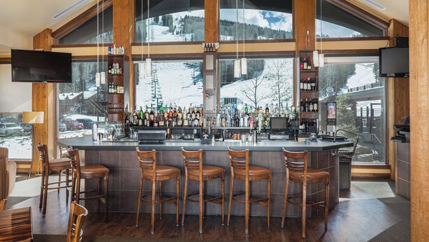 Een bar met een groot raamzicht op een besneeuwde berg, houten barstoelen en diverse flessen en glazen op de planken.