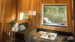 Interior acogedor de una cabaña con paredes de madera, un sofá, una mesa de centro y un ventanal con vistas a los árboles.