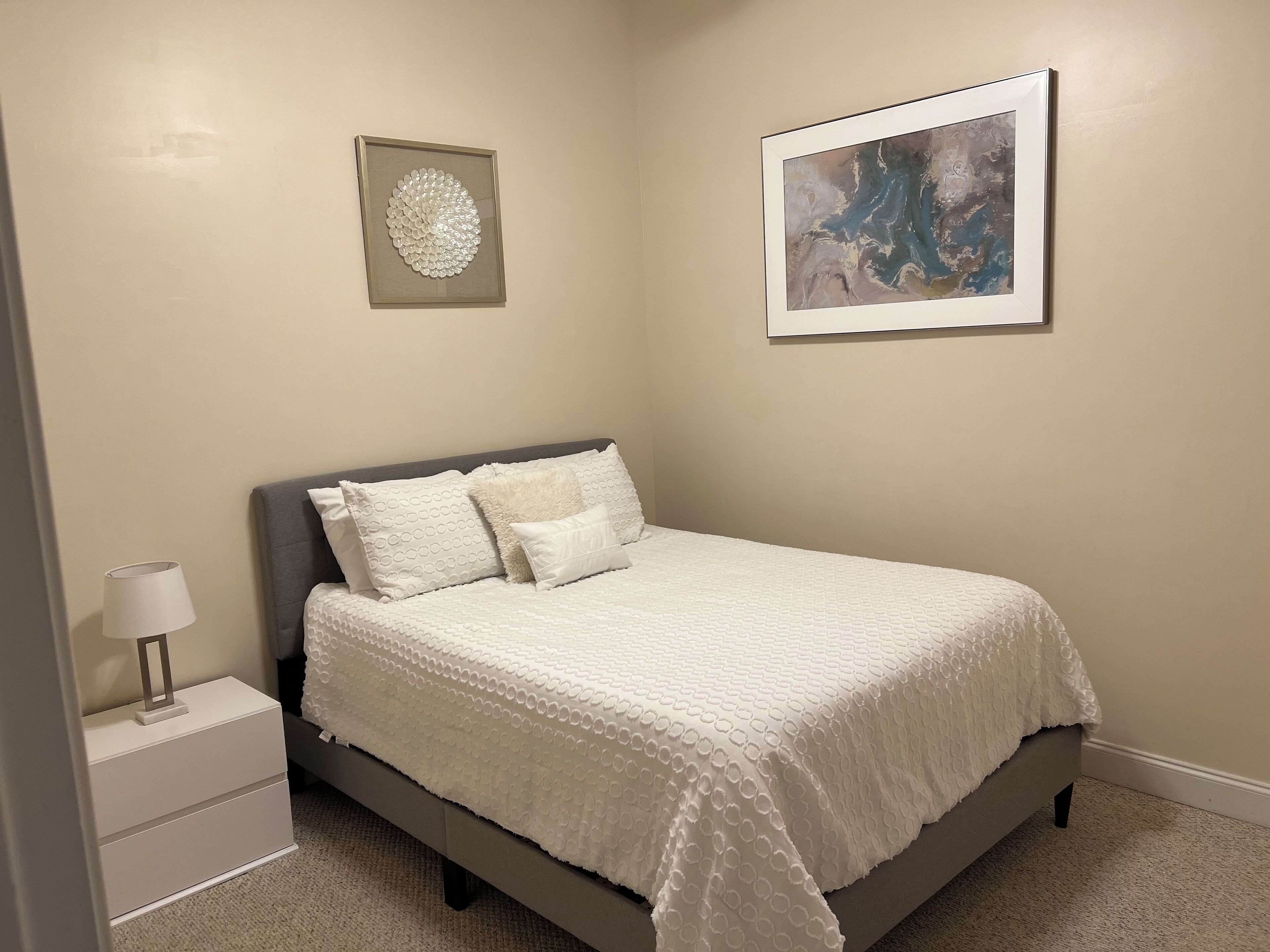 A bedroom with a bed, nightstand, lamp, and two framed artworks on the walls.